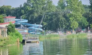 Letnia zabawa na **zalewie zemborzyckim**: niebieskie zjeżdżalnie wodne, ludzie kąpiący się i relaksujący się na plaży, otoczeni zielenią.