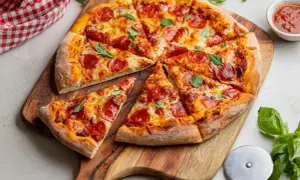 Pokrojona pizza pepperoni z serem i bazylią na desce, obok sos i nóż do pizzy.