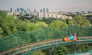 Dwie osoby zjeżdżają na torze saneczkowym na tle panoramy Warszawy. Wspaniała atrakcja dla każdego, kto chce spędzić czas w stolicy z dzieckiem.