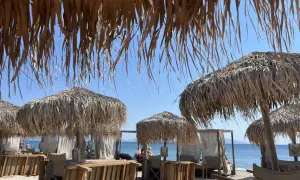 Słomiane parasole i leżaki na plaży Tigaki, z widokiem na spokojne, błękitne morze i bezchmurne niebo.