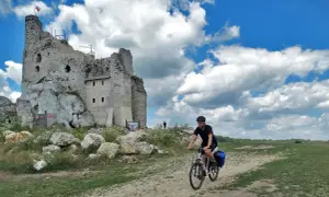 Rowerzysta przemierza malownicze **Jura Krakowsko-Częstochowska szlaki**, mijając ruiny zamku na skale.