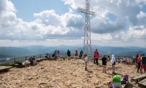 Ludzie odpoczywają na szczycie, podziwiając widoki na Bieszczady. Metalowy krzyż góruje nad kamienistym terenem, a w oddali widać łagodne pasma górskie.