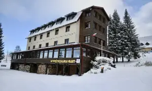 Zimowy hotel z drewnianymi elementami, idealny na zieleniec noclegi blisko stoku. Wokół śnieg i wysokie choinki.