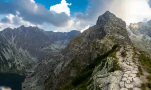 Kamienista ścieżka prowadzi na szczyt w Tatrach Zachodnich. W dole jezioro, w tle skaliste pasmo górskie pod błękitnym niebem z chmurami.
