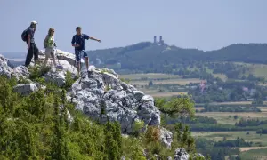 Trzech turystów na skale podziwia krajobraz Gór Świętokrzyskich. Jeden z nich wskazuje kierunek, gdzie w oddali widać ruiny zamku.