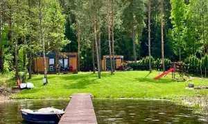 Drewniany pomost prowadzi do jeziora, gdzie cumuje łódka. Na brzegu, wśród drzew, stoją domki letniskowe, plac zabaw i rowery. Idealne miejsce na wypoczynek w **kiekrz**.