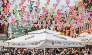 Kolorowe chorągiewki ozdabiają ulicę podczas Jarmarku Świętego Dominika. Ludzie spacerują pod białymi parasolami z napisami "Jarmark Św. Dominika" i "Saint Dominic's Fair".