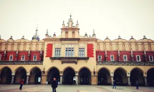 Sukiennice na Rynku Głównym w Krakowie. Kilka osób spaceruje po pustym placu przed zabytkowym budynkiem z arkadami.