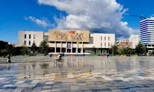 Plac w Tiranie, stolicy Albanii, z monumentalnym budynkiem i mozaiką na fasadzie. Na pierwszym planie mokra nawierzchnia odbija niebo.