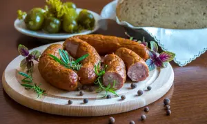 Kiełbasa podwawelska na desce z rozmarynem, obok ogórki i chleb.