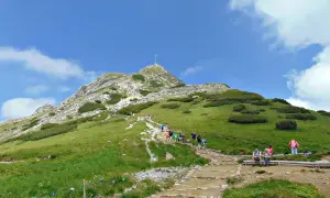 Tłum turystów wspina się kamienistym szlakiem na Giewont. Na szczycie widoczny krzyż.