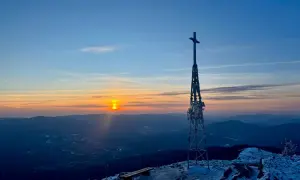 Zachód słońca nad górami, wieża z krzyżem na szczycie, ławki pokryte śniegiem. W oddali widać lasy, w tym smerek.