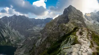 Kamienista ścieżka prowadzi na szczyt w Tatrach Zachodnich. W dole jezioro, w tle skaliste pasmo górskie pod błękitnym niebem z chmurami.