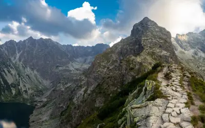 Kamienista ścieżka prowadzi na szczyt w Tatrach Zachodnich. W dole jezioro, w tle skaliste pasmo górskie pod błękitnym niebem z chmurami.