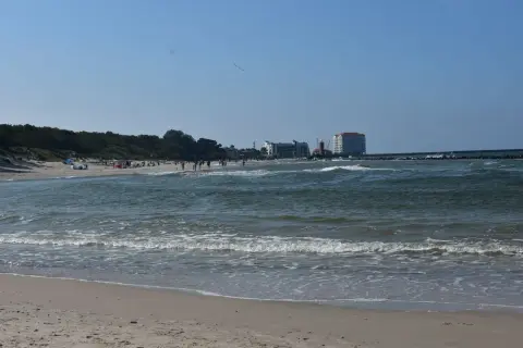 Słoneczny dzień nad Bałtykiem, plaża pełna ludzi, a w oddali widać miasto u ujścia Wieprzy.