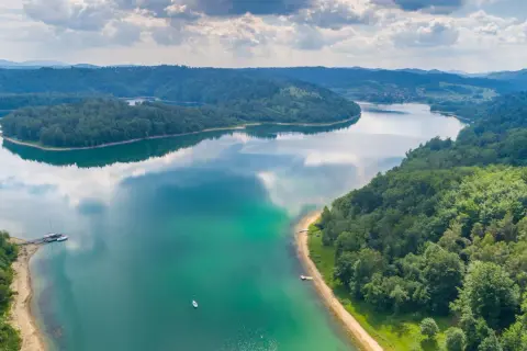 Malownicze pojezierza z zielonymi lasami i turkusową wodą. Na brzegu cumują żaglówki, a na tafli jeziora pływa mała łódka.