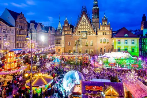 Tłoczny jarmark we Wrocławiu nocą, rozświetlony tysiącami lampek. Widać Ratusz, karuzelę i stragany.