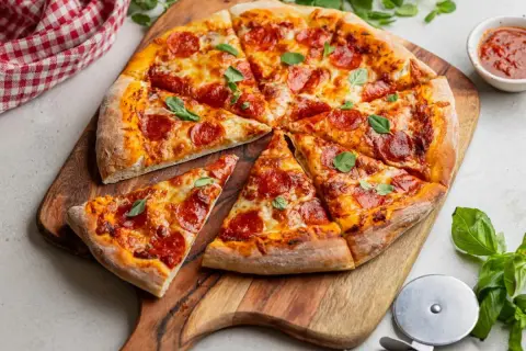 Pokrojona pizza pepperoni z serem i bazylią na desce, obok sos i nóż do pizzy.