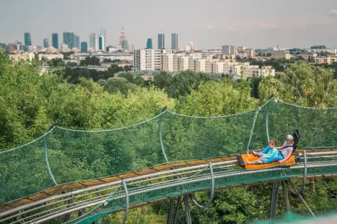 Dwie osoby zjeżdżają na torze saneczkowym na tle panoramy Warszawy. Wspaniała atrakcja dla każdego, kto chce spędzić czas w stolicy z dzieckiem.