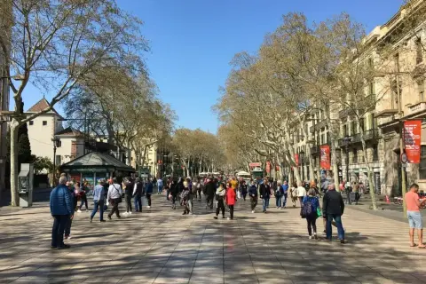 Tłum ludzi spaceruje po słynnej alei La Rambla w Barcelonie, otoczonej drzewami i zabytkowymi budynkami.