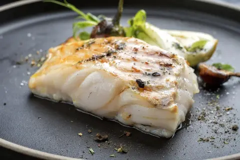 Usmażony filet ryby miruna z ziołami i warzywami na czarnym talerzu.
