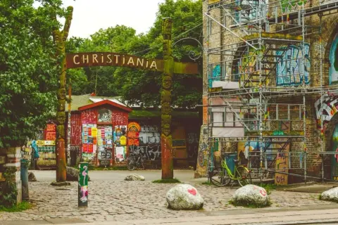 Drewniany totem i tablica z napisem "CHRISTIANIA" wita w tej wolnej dzielnicy. Obok czerwony budynek z plakatami i rowery.