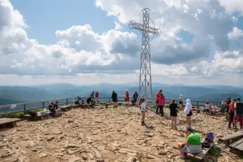Ludzie odpoczywają na szczycie, podziwiając widoki na Bieszczady. Metalowy krzyż góruje nad kamienistym terenem, a w oddali widać łagodne pasma górskie.