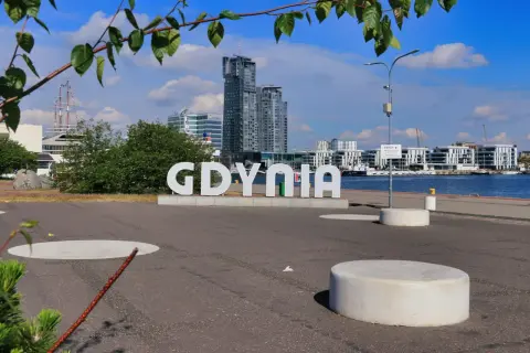 Napis "GDYNIA" na tle nowoczesnych budynków i żaglowca. Widok na port, idealny na spacer po gdynia stare miasto.