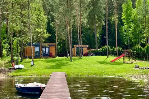 Drewniany pomost prowadzi do jeziora, gdzie cumuje łódka. Na brzegu, wśród drzew, stoją domki letniskowe, plac zabaw i rowery. Idealne miejsce na wypoczynek w **kiekrz**.