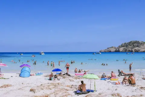 Słoneczna plaża Cala Agulla tętni życiem. Ludzie kąpią się w krystalicznie czystej wodzie, opalają na piasku i odpoczywają pod parasolami.