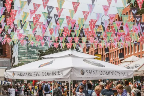 Kolorowe chorągiewki ozdabiają ulicę podczas Jarmarku Świętego Dominika. Ludzie spacerują pod białymi parasolami z napisami "Jarmark Św. Dominika" i "Saint Dominic's Fair".