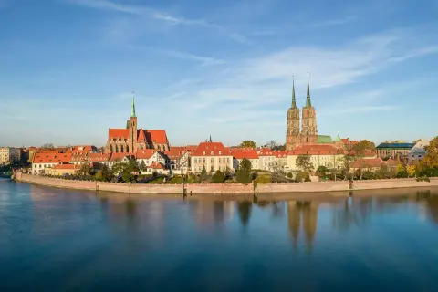 Widok na Ostrów Tumski, serce wrocławskiej starówki, z katedrą i innymi zabytkowymi budynkami odbijającymi się w spokojnej wodzie.