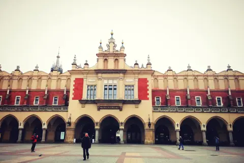 Sukiennice na Rynku Głównym w Krakowie. Kilka osób spaceruje po pustym placu przed zabytkowym budynkiem z arkadami.
