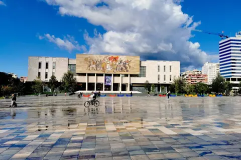 Plac w Tiranie, stolicy Albanii, z monumentalnym budynkiem i mozaiką na fasadzie. Na pierwszym planie mokra nawierzchnia odbija niebo.