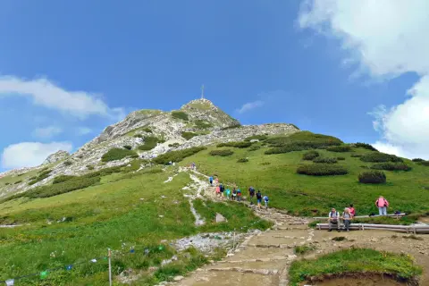 Tłum turystów wspina się kamienistym szlakiem na Giewont. Na szczycie widoczny krzyż.