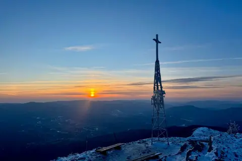 Zachód słońca nad górami, wieża z krzyżem na szczycie, ławki pokryte śniegiem. W oddali widać lasy, w tym smerek.