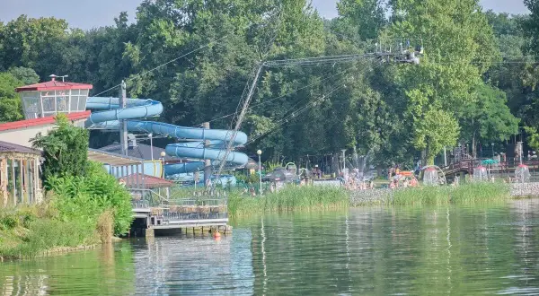 Letnia zabawa na **zalewie zemborzyckim**: niebieskie zjeżdżalnie wodne, ludzie kąpiący się i relaksujący się na plaży, otoczeni zielenią.