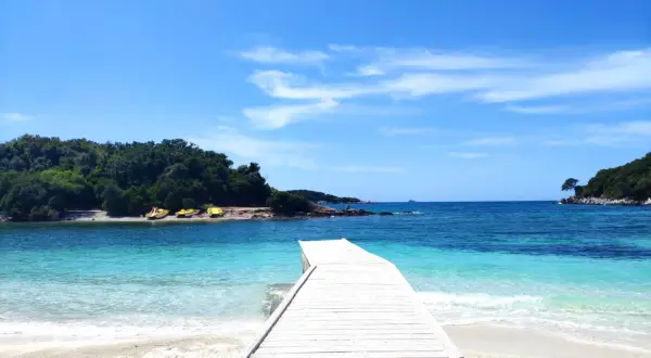 Białe molo prowadzi do krystalicznie czystej wody na Ksamil Beach. W oddali zielone wyspy i błękitne niebo.