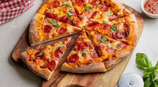 Pokrojona pizza pepperoni z serem i bazylią na desce, obok sos i nóż do pizzy.