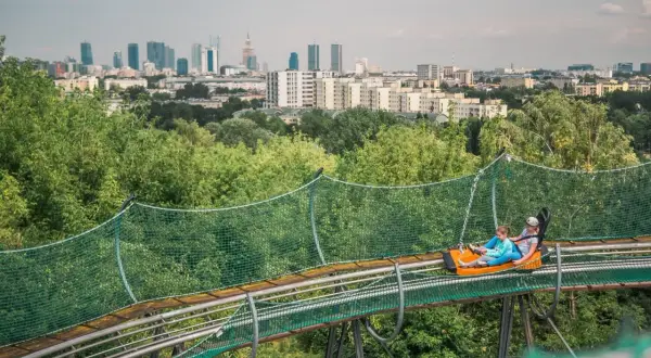 Dwie osoby zjeżdżają na torze saneczkowym na tle panoramy Warszawy. Wspaniała atrakcja dla każdego, kto chce spędzić czas w stolicy z dzieckiem.