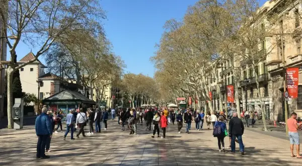 Tłum ludzi spaceruje po słynnej alei La Rambla w Barcelonie, otoczonej drzewami i zabytkowymi budynkami.