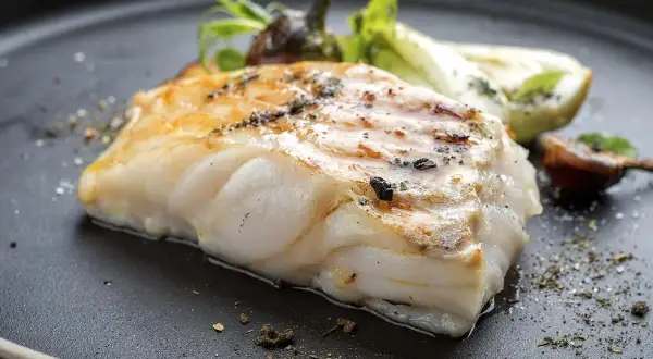 Usmażony filet ryby miruna z ziołami i warzywami na czarnym talerzu.