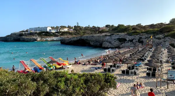 Słoneczna plaża Cala Domingos tętni życiem. Woda jest turkusowa, a na piasku widać rzędy parasoli i leżaków. Dzieci bawią się na kolorowych zjeżdżalniach.