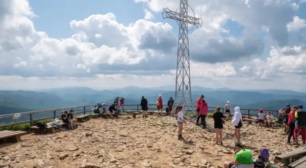 Ludzie odpoczywają na szczycie, podziwiając widoki na Bieszczady. Metalowy krzyż góruje nad kamienistym terenem, a w oddali widać łagodne pasma górskie.