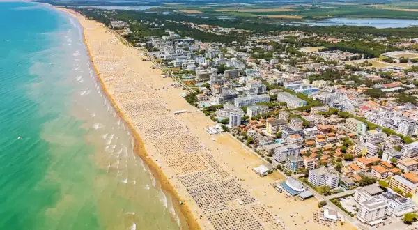 Słoneczna bibione plaża z tysiącami leżaków i parasoli, turkusowe morze i miasto w tle.