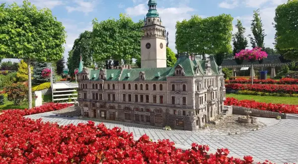Miniatura ratusza z wieżą zegarową, otoczona czerwonymi kwiatami, w miniaturowej kotlinie kłodzkiej.