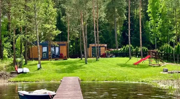 Drewniany pomost prowadzi do jeziora, gdzie cumuje łódka. Na brzegu, wśród drzew, stoją domki letniskowe, plac zabaw i rowery. Idealne miejsce na wypoczynek w **kiekrz**.