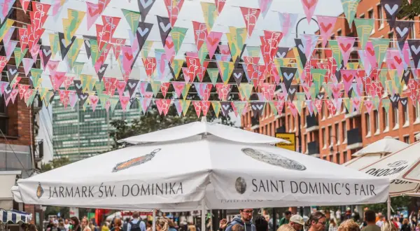 Kolorowe chorągiewki ozdabiają ulicę podczas Jarmarku Świętego Dominika. Ludzie spacerują pod białymi parasolami z napisami "Jarmark Św. Dominika" i "Saint Dominic's Fair".