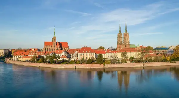 Widok na Ostrów Tumski, serce wrocławskiej starówki, z katedrą i innymi zabytkowymi budynkami odbijającymi się w spokojnej wodzie.