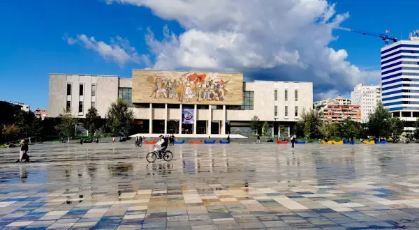 Plac w Tiranie, stolicy Albanii, z monumentalnym budynkiem i mozaiką na fasadzie. Na pierwszym planie mokra nawierzchnia odbija niebo.